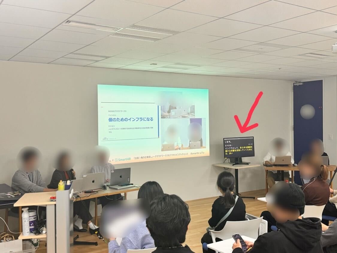 写真:アクセシビリティイベント会場の様子。中央のスクリーンにはクラウドワークスのスライドが投影されており、右側の小型モニターにはUDトークによるリアルタイム字幕が表示されています。参加者が椅子に座り、講演を見守っている。