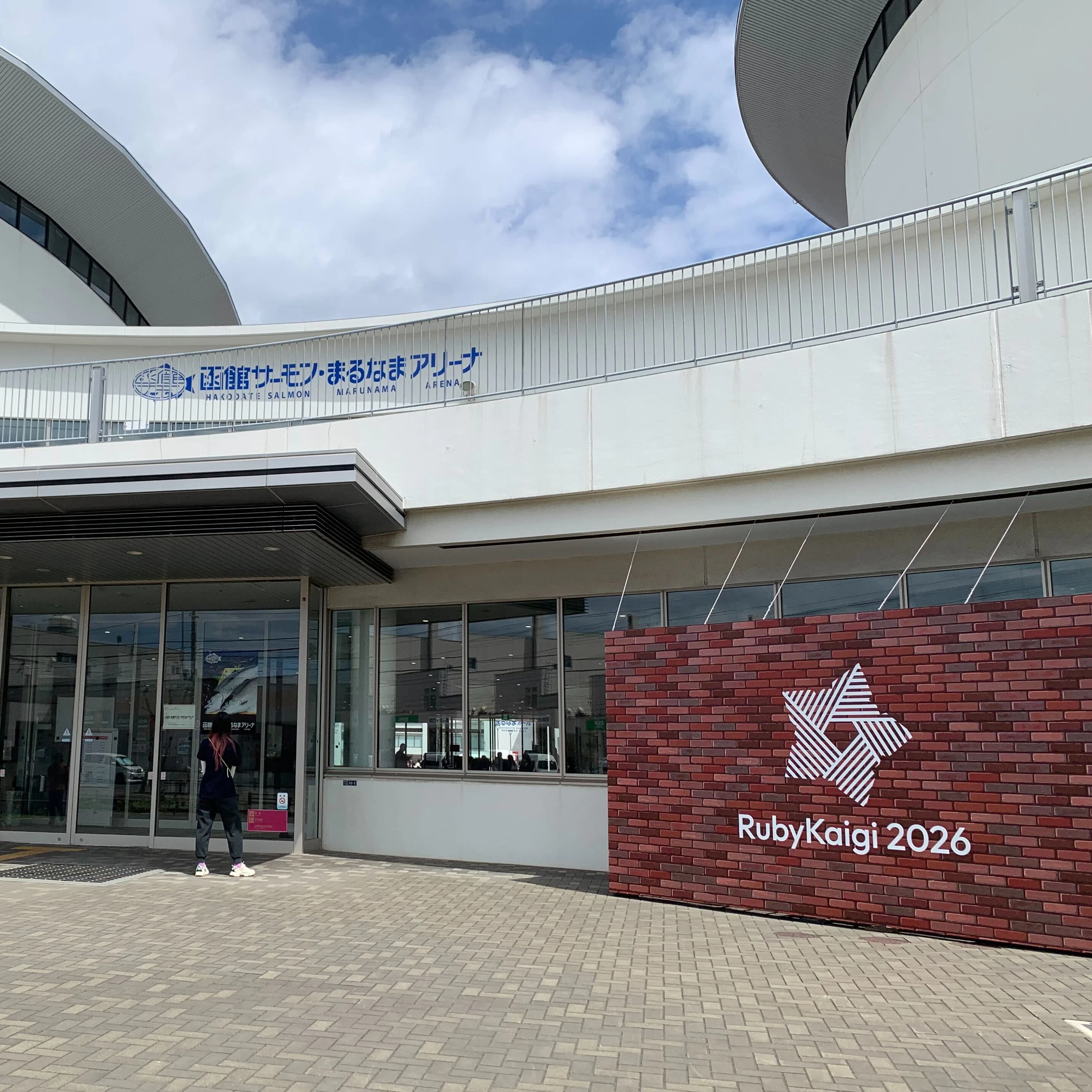 RubyKaigi 2026の会場「函館サーモン・まるなまアリーナ」正面の様子。レンガのデザインの立て看板が掲示された入口が見える。.jpg