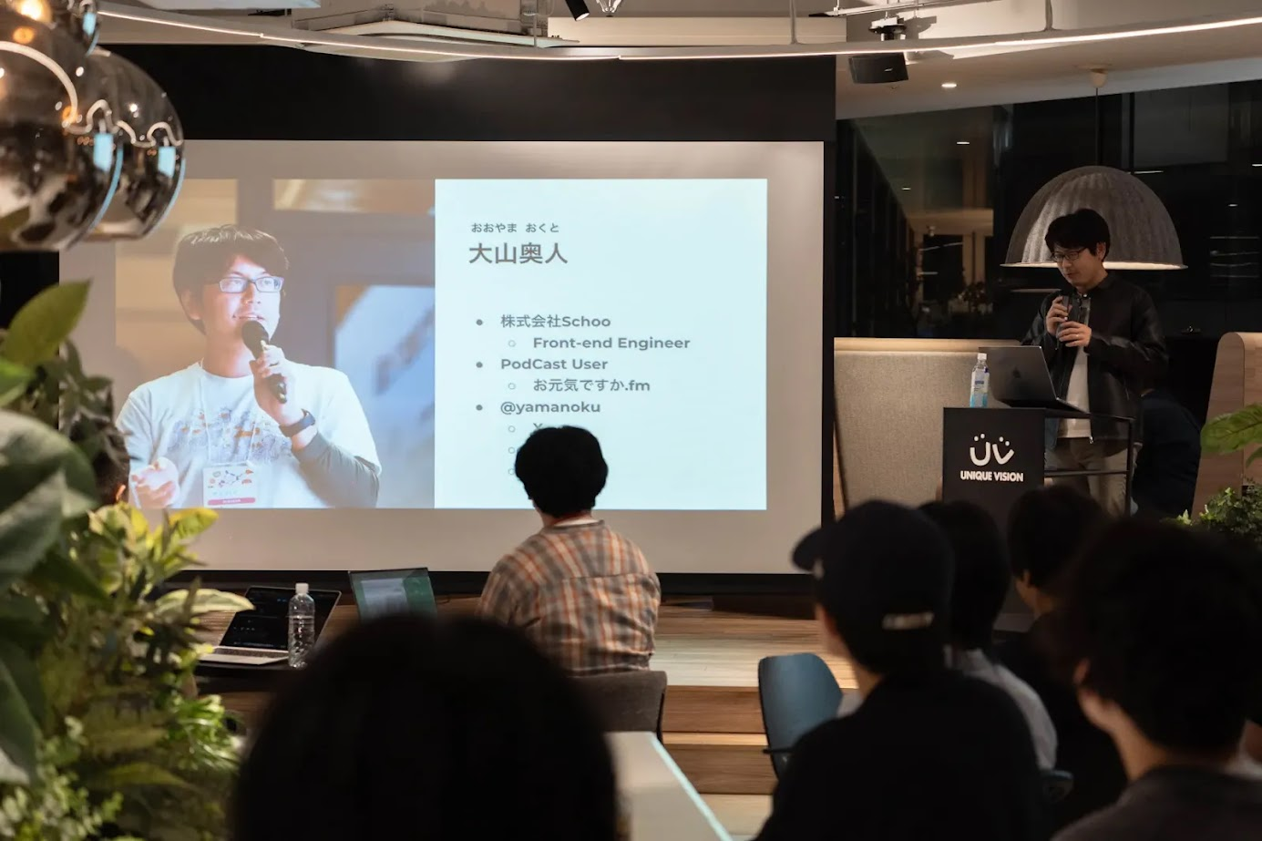 プレゼンテーションを行う大山と、スクリーンを見ている聴衆。スクリーンには大山奥人の顔写真と名前、所属、ポッドキャストの情報、SNSアカウントが記載されている。