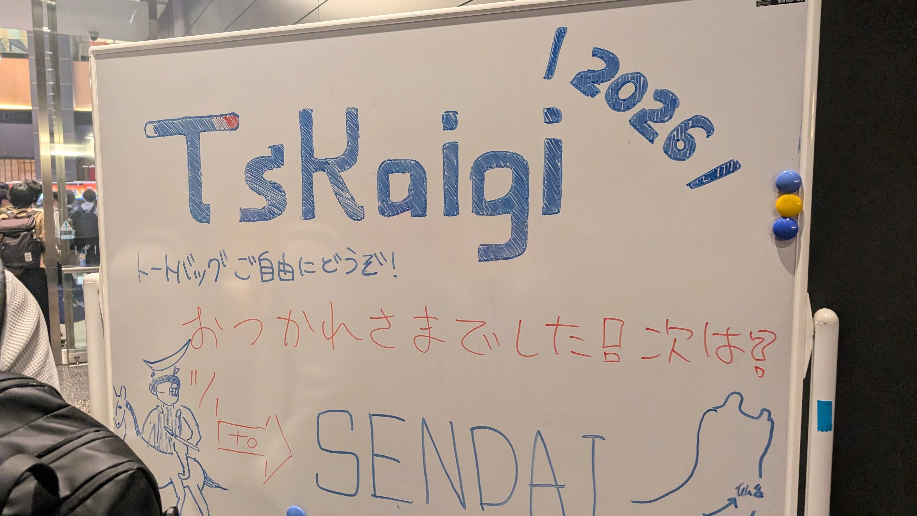 TSKaigi 2026の会場出口に設置されたホワイトボード。青と赤の手書き文字で「TskKaigi 2026」「トートバッグご自由にどうぞ!」「おつかれさまでした!次は?」などのメッセージがあり、伊達政宗像のイラストと「SENDAl」 の文字、日本地図の描き込みがある。
