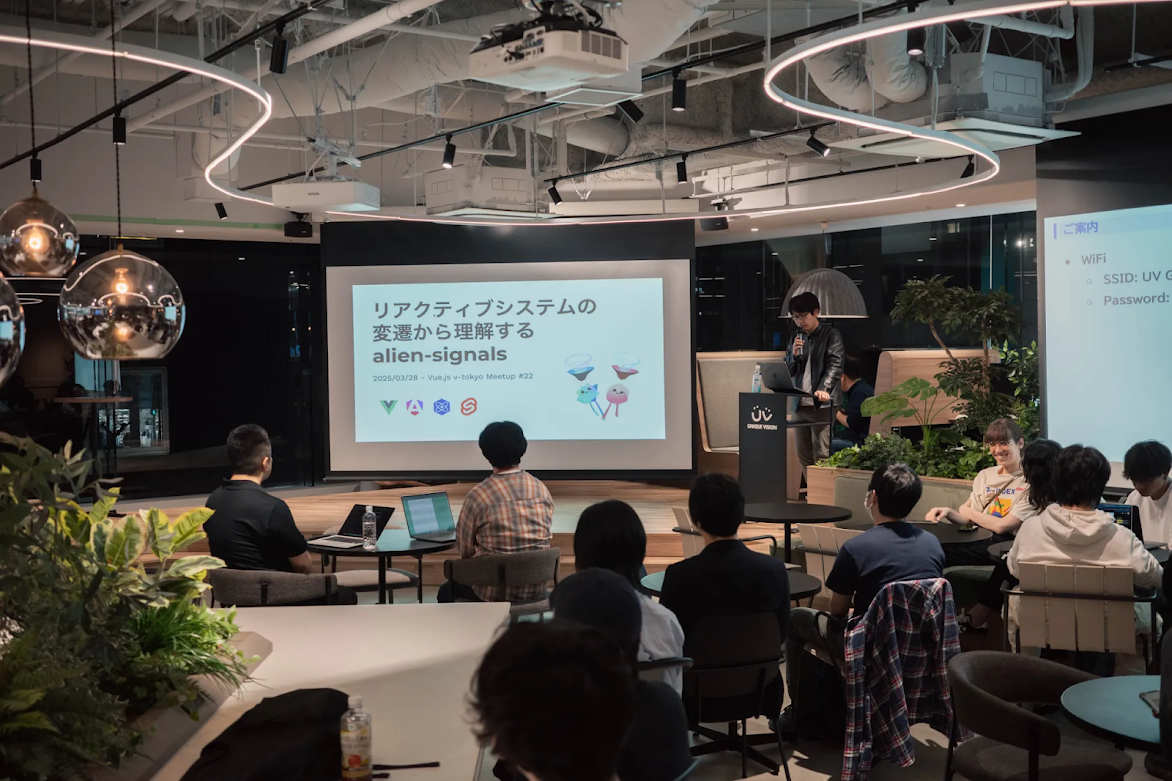 当日の勉強会会場でもあるユニークビジョン社で登壇・発表する大山の様子。登壇スライドの表紙が映し出されている。周囲には椅子に座っている聴衆が見える。室内には黒いテーブルと椅子が並べられており、天井の照明は白色で、部屋全体は白色、天井の一部も白色で覆われている。