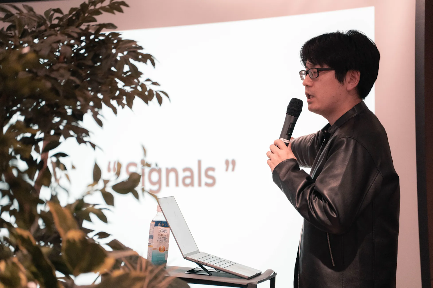 マイクを持ち話をする大山奥人の上半身の画像。スクリーンには大きく「signals」という文字が表示されている。左側には観葉植物が配置され、手前の机の上にはノートパソコンと飲み物が置かれている。